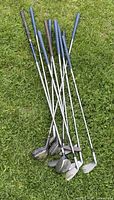 Seven golf clubs laid on grass showing shafts and rubber grips mostly in blue and black colors