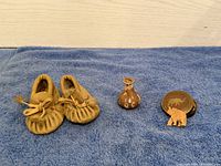 Photo showing the full small item lot on a blue cloth: baby moccasins, pottery vase, carved wooden elephant, and tin lid with elephant design.