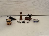Photo shows a collection of miniature collectibles on a wooden surface including a wooden cat figurine, miniature sewing machine, small cannon, ceramic cup, ceramic basket, and three tiny animal figurines.