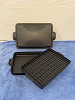 Pampered Chef microwaveable bacon pan unassembled showing cover, slotted grilling rack and drip tray on a blue cloth surface against light wood background.