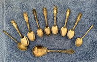 Top view of all 11 spoons arranged side by side on a blue towel, showing ornate decorative handles and varying bowl shapes