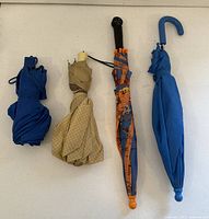 Four folded umbrellas laid out on a surface in a row: navy blue compact, beige with polka dots, colorful kids umbrella, and bright blue kids umbrella with curved handle.