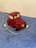 Red ceramic lamp in the shape of a vintage car, showing the front and sides with painted details and electrical cord.