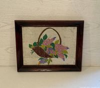 Vintage framed embroidery of a floral basket with purple and yellow flowers, green leaves, and a brown basket, placed against a light wood background.