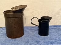 Pair of vintage metal measuring cans, larger rusted metal can with spout on left, smaller black metal can with handle on right, displayed on blue cloth against wooden background.
