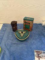 Three vintage tins placed on a blue towel: Red Rose tea tin (rectangular), a cigar tin (cylindrical), and a heart-shaped decorative tin.