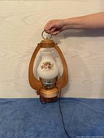 Full view of the lantern light held by hand showing natural wood frame and floral glass shade with electric cord.