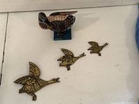 Front view of three brass flying geese arranged in descending size from left to right, and one painted wooden goose on blue base behind them.