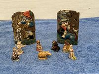 Two carved child figurines on wood blocks shown with seven miniature animal and character figurines on blue cloth.