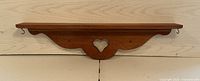 Front view of a wooden wall shelf with heart-shaped decorative cut-out and two hooks below the shelf surface.