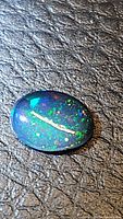 Close-up photo of an oval black opal gemstone with bright multicolored flashes on a textured dark surface.