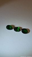 Three oval cut tsavorite garnet stones displayed on a white surface, showing dark vivid green color and transparency.