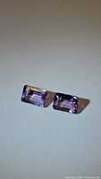 Two rectangular emerald-cut light purple amethyst gemstones set next to each other on a plain neutral background, showing matching size and clarity.