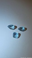 Photo showing three natural blue aquamarine gemstones on a white background, highlighting pear and cushion cuts.