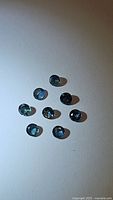 Eight small natural round cut blue sapphires with teal green undertones spaced on a white background showing their size and color variation.