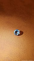 Round cut natural Brazilian aquamarine gemstone, approximately 6.7mm diameter shown from above on tan background.