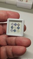 Natural Ceylon sapphires shown in small white display case, held in hand for scale, showing 12 rectangular calibrated stones in shades of blue and near colorless.