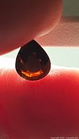 Pear-shaped deep orange tourmaline held between fingers, light shining through to show color and transparency.