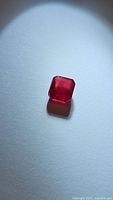 Close-up image of a deep red square cut ruby gemstone on a light background.