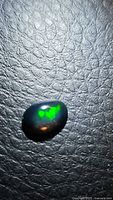 Close-up photo of the oval black opal gemstone showing green play-of-color on a textured black surface.