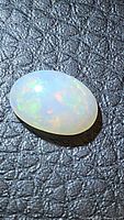 Close-up photo of an oval cabochon natural Ethiopian opal gemstone showing multi-color flash on textured black surface.