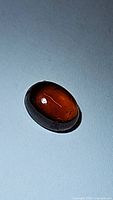 Oval cabochon cut burnt orange hessonite garnet gemstone photographed on a plain surface with visible light reflections.