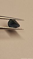 Close-up view of pear-shaped blue sapphire held by tweezers showing deep blue color and green flashes.