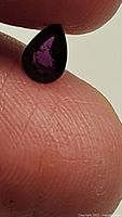 Close-up photo of a pear-shaped, faceted dark orange fire opal gemstone placed on the tip of a finger, showing internal flashes of purple and other colors.
