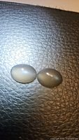 Two oval cabochon natural moonstones of smokey platinum color, placed side-by-side on a textured dark surface, showing size and color match.