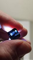 Close-up of square cut blue purple tourmaline gemstone held between thumb and finger, illuminated to show transparency and color variation.