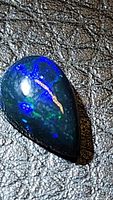 Close-up photo of a pear-shaped natural Ethiopian black opal cabochon showing electric blue and green rolling flash colors on a dark base.