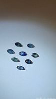 Nine natural marquis cut sapphires in various shades of blue and green arranged on a white surface.