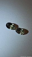 Two natural platinum green untreated sapphires placed side by side on a white surface under lighting showing their oval cut and internal clarity.