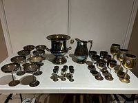 Wide shot of silver plated champagne bucket, pitcher, and more than 20 goblets of varying sizes and shapes arranged on a white folding table against a plain wall.