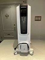 Front view of white Delonghi radiant heater with digital display and controls, power cord wrapped on front bottom, on caster wheels.