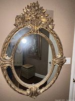 Full view of gold framed oval mirror with ornate floral decorative top and beaded trim around the edge.