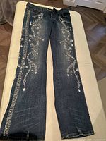 Full length view of dark wash denim jeans laid flat, showing the sequin decorated swirling patterns on the legs.