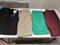 Five pairs of ladies dress pants, colors black, dark brown with embroidery, beige, green lace, and burgundy, folded on table.