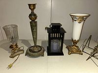 Four items on a table including a clear glass electric vase with gold-tone base, a tall greenish metal candlestick, a black metal lantern with candle, and a tall white and gold vase.