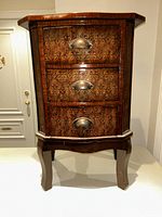 Front view of the wooden nightstand showing all three drawers with decorative metal cup handles and a carved apron under the drawers.