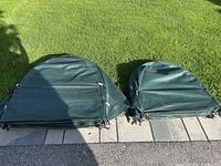 Shows two piles of folded shrub covers on a paved edge next to grass; the pile on the left is larger and taller covers, the pile on the right smaller and shorter covers.