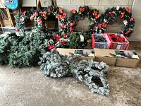 Photo showing four green 4-foot wreaths decorated with red poinsettia flowers, cream flowers, and pinecones, two boxed frosted artificial Christmas trees, and bins containing string lights and decorations.