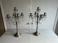 Front view of two silver plated candelabras standing on a flat surface against a white background. Both have multiple clear hanging crystal drops and five candle holders.