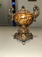 Front view of the decorative small urn showing mottled brown finish, ornate handles, and detailed base.
