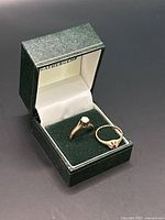Two yellow gold rings (white and red stones) displayed in green ring box with lid open