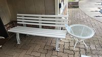 White outdoor bench with plastic-coated slats and white metal legs, set on outdoor paving.