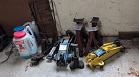 Photo showing four red jack stands, two hydraulic floor jacks (yellow and blue), various hitches, plastic jug of ice melt, and set of ice cleats in a garage.