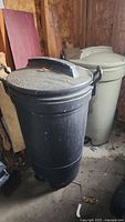 Side view of two garbage cans in wooden shed, showing the black Gott can in front and beige Rubbermaid can behind.