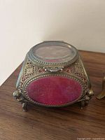 Angled front view of gold filigree ormolu jewellery box showing detailed filigree and red velvet panel inside, standing on cherub feet.