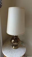 Front angle of table lamp showing brass-colored spherical base and off-white linen style cylindrical shade with visible loose piping.
